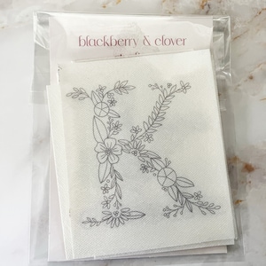May include: A white square fabric sheet featuring a gray line art illustration of the letter K formed by various floral and leaf patterns. The sheet is packaged in a clear plastic sleeve with a card labeled blackberry & clover at the top.