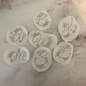 May include: Seven white paper circles with gray floral designs and a single letter in each circle. The letters are A, W, M, S, E, C, and K.