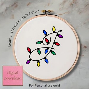 May include: An embroidery hoop containing white fabric with a black line art design of a string of Christmas lights shaped like the letter J. The lights are colored red, blue, green, yellow, and purple. Text reads Letter J - 4" Christmas Light Pattern.