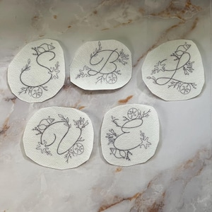 May include: Five white fabric patches with embroidered gray letters: S, B, L, D, and E. Each letter is decorated with seashells, starfish, and botanical elements, creating a coastal theme. The patches are arranged on a marble-patterned surface.