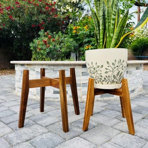 May include: Two wooden plant stands with a mid-century modern design. One stand holds a cream-colored ceramic pot with a green leaf pattern. The other stand is empty. The stands are made of dark brown wood and are on a stone patio.