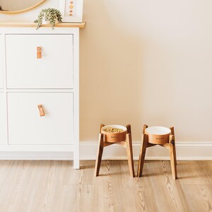May include: Two elevated pet bowls on wooden stands. The left bowl is tan with the text "NOSH" and contains dry food. The right bowl is tan with the text "SLURP" and contains water. Both bowls have white interiors and are supported by four-legged wooden frames.