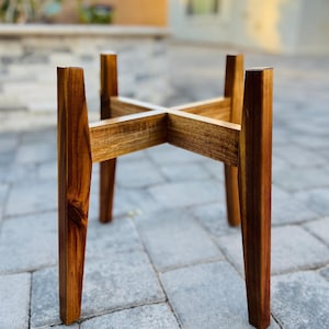 May include: A wooden plant stand with a cross-shaped top and four angled legs. The stand is made of a warm-toned wood, showcasing the natural grain. The design is simple and modern, suitable for displaying potted plants.
