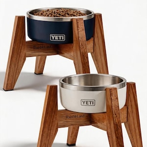 May include: Two elevated pet bowls featuring stainless steel interiors and coloured exteriors, one navy blue and one white, each branded with the YETI logo. The bowls are held by wooden stands labelled RainbleLand with four angled legs and black feet.