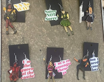 May include: Six pairs of earrings featuring characters and quotes from the musical Six. The earrings are made of acrylic and feature colorful designs. The quotes on the earrings include "I'm the queen of the castle!" and "I'm a 10 amongst these 38".