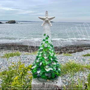 May include: A decorative Christmas tree made of sea glass, with a white starfish topper. The tree is composed of green and white sea glass pieces, standing on a wooden base. The background shows a beach and ocean.