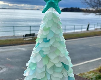 May include: A turquoise sea glass Christmas tree with a turquoise starfish on top. The tree is made of many small pieces of sea glass in shades of blue and green. The tree is sitting on a wooden surface.