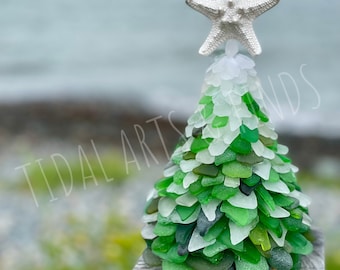 May include: A handcrafted Christmas tree made of sea glass in shades of green and white, topped with a silver starfish. The tree is displayed on a wooden post with a blurred ocean background. The text "TIDAL ARTS & SANDS" is visible.