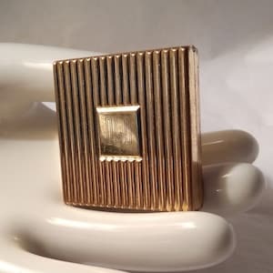 May include: A gold-colored compact with a ribbed design and the letters "D.G.D." embossed in the center.