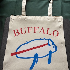 Può includere: Borsa tote color crema con la scritta "BUFFALO" in lettere rosse. Sotto, una grafica di bufalo bianco con una striscia rossa e dettagli blu. La borsa ha manici color crema.