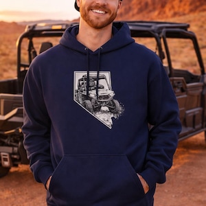 May include: Navy blue hoodie featuring a black and white graphic of an off-road vehicle within a Nevada state outline. The hoodie has a drawstring hood and a front pocket. The person is wearing a black baseball cap and blue jeans.