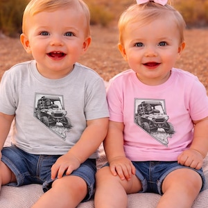 May include: Two babies wearing matching t-shirts and denim shorts. One shirt is light gray, the other is pink. Each shirt features a black and white graphic of an off-road vehicle inside a Nevada state outline.