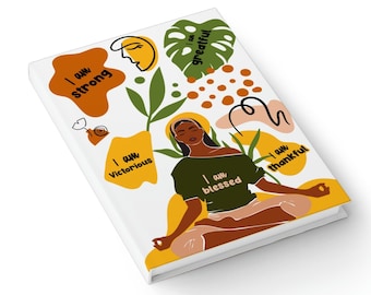 Op de afbeelding: Een wit dagboek met een kleurrijk ontwerp met een mediterende vrouw met de woorden "Ik ben sterk", "Ik ben overwinnend", "Ik ben gezegend", "Ik ben dankbaar" en "Ik ben dankbaar" in zwarte tekst.