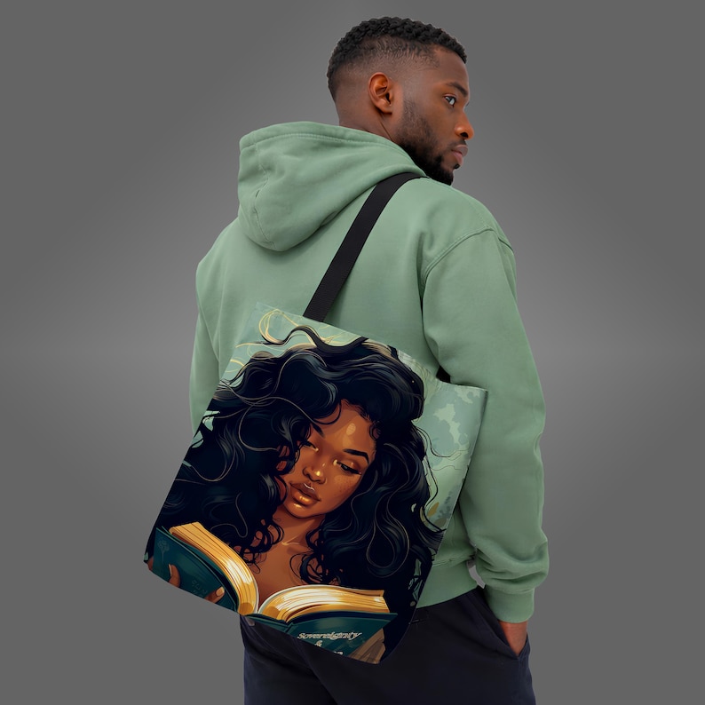 May include: A tote bag featuring an illustration of a Black woman with long, dark, curly hair reading a book. The book has gold-colored pages and the word "Sovereignty" is visible. The bag has a black strap and is carried over the shoulder.