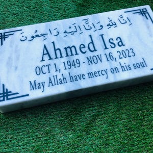 Puede incluir: Placa conmemorativa rectangular de mármol con el nombre "Ahmed Isa", fechas de nacimiento y defunción, y una inscripción religiosa. La placa tiene diseños decorativos en las esquinas y está sobre un fondo verde.