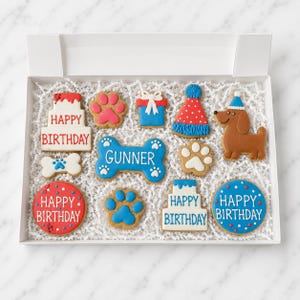 May include: A box of decorated dog-themed cookies. The cookies include a bone, paw prints, a gift, a party hat, a dachshund, and cookies that say "Happy Birthday". The cookie colors are red, white, blue, and brown.