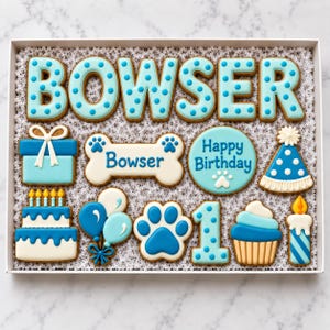 May include: A collection of blue and white decorated sugar cookies in a box. The cookies include the name BOWSER in polka-dot letters, a gift box, a bone with the name Bowser, a round Happy Birthday cookie, a party hat, a cake, balloons, a paw print, the number 1, a cupcake, and a candle.