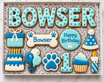 May include: A collection of blue and white decorated sugar cookies in a box. The cookies include the name BOWSER in polka-dot letters, a gift box, a bone with the name Bowser, a round Happy Birthday cookie, a party hat, a cake, balloons, a paw print, the number 1, a cupcake, and a candle.