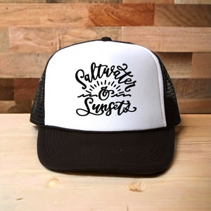 May include: A black and white trucker hat with a white front panel and a black mesh back. The front panel features a black graphic that says "Saltwater & Sunsets".