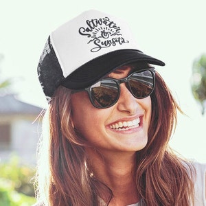 May include: A white and black trucker hat with the text "Saltwater & Sunsets" in black script. The hat has a black mesh back and a white front panel. The person wearing the hat is smiling and wearing sunglasses.