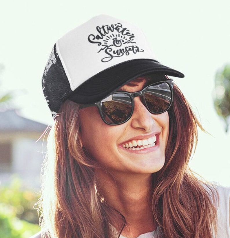May include: A white and black trucker hat with the text "Saltwater & Sunsets" in black script. The hat has a black mesh back and a white front panel. The person wearing the hat is smiling and wearing sunglasses.