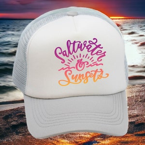 May include: White trucker hat with a mesh back. The front of the hat has a pink, orange, and yellow graphic that says "Saltwater & Sunsets".