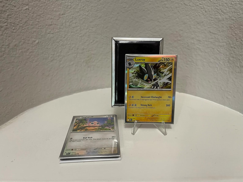 Puede incluir: Dos cartas coleccionables de Pok&eacute;mon, una con Luxray y otra con un Jigglypuff. La carta de Luxray es amarilla y negra, expuesta en un soporte transparente. Un espejo con marco negro est&aacute; detr&aacute;s de la carta de Luxray. La carta de Jigglypuff es plateada y rosa.