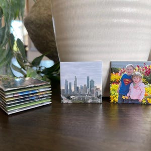 Puede incluir: Una pila de impresiones fotogr&aacute;ficas cuadradas y dos impresiones cuadradas individuales. Una impresi&oacute;n muestra un horizonte urbano, y la otra muestra a dos ni&ntilde;os en un campo de flores. Las impresiones est&aacute;n sobre una superficie marr&oacute;n oscuro.