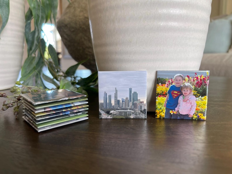 Puede incluir: Una pila de impresiones fotogr&aacute;ficas cuadradas y dos impresiones cuadradas individuales. Una impresi&oacute;n muestra un horizonte urbano, y la otra muestra a dos ni&ntilde;os en un campo de flores. Las impresiones est&aacute;n sobre una superficie marr&oacute;n oscuro.