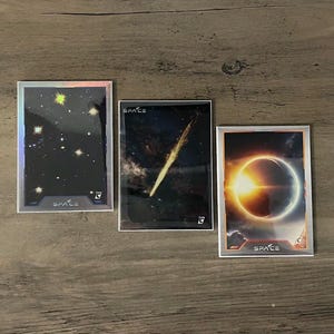 Puede incluir: Tres cromos con im&aacute;genes espaciales. Uno muestra un cielo estrellado, otro un cometa y el tercero un eclipse solar. Cada cromo tiene la palabra "SPACE" y un n&uacute;mero.