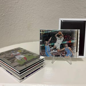 Puede incluir: Un conjunto de tarjetas coleccionables deportivas con jugadores de f&uacute;tbol americano y baloncesto. Una tarjeta se muestra en un soporte acr&iacute;lico transparente, mostrando a un jugador de baloncesto. Tambi&eacute;n se ve un im&aacute;n enmarcado blanco.