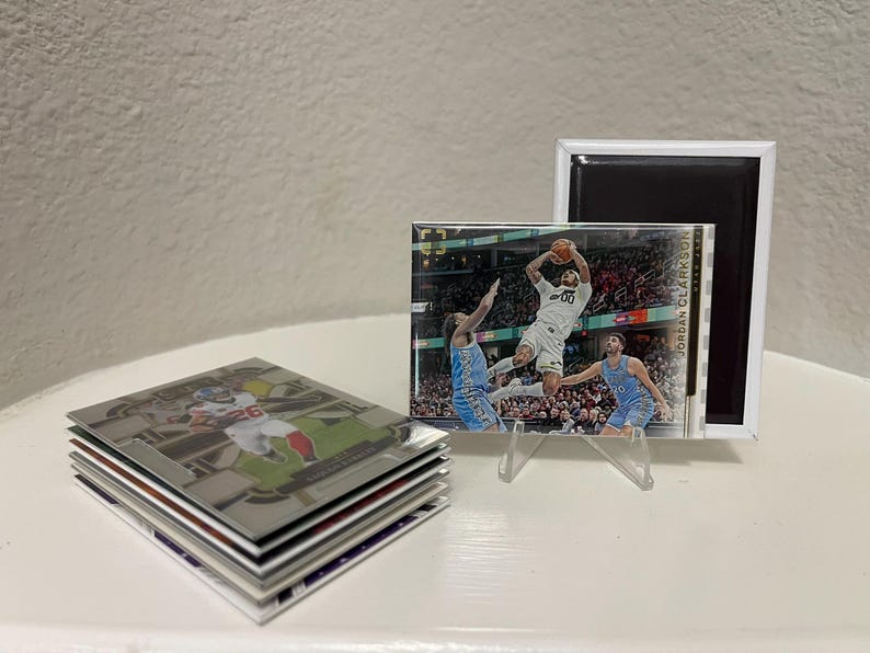 Puede incluir: Un conjunto de tarjetas coleccionables deportivas con jugadores de f&uacute;tbol americano y baloncesto. Una tarjeta se muestra en un soporte acr&iacute;lico transparente, mostrando a un jugador de baloncesto. Tambi&eacute;n se ve un im&aacute;n enmarcado blanco.