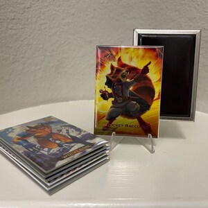 Puede incluir: Una pila de cromos con Wolverine y una tarjeta de Rocket Raccoon en exhibici&oacute;n. La tarjeta de Rocket Raccoon est&aacute; en un soporte transparente, con un marco magn&eacute;tico negro detr&aacute;s. La tarjeta de Rocket Raccoon tiene un fondo ardiente.