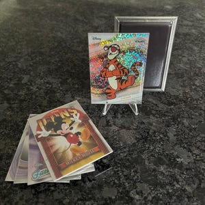 Puede incluir: Tarjetas coleccionables de Disney con personajes como Tigger y Mickey Mouse. La tarjeta de Tigger se muestra en un soporte transparente, con una pila de tarjetas cerca. Tambi&eacute;n hay un marco magn&eacute;tico plateado.
