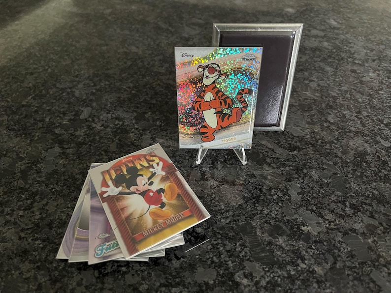 Puede incluir: Tarjetas coleccionables de Disney con personajes como Tigger y Mickey Mouse. La tarjeta de Tigger se muestra en un soporte transparente, con una pila de tarjetas cerca. Tambi&eacute;n hay un marco magn&eacute;tico plateado.