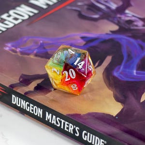 May include: A colorful, multi-sided die with the number 20 prominently displayed, resting on a Dungeons & Dragons Dungeon Master's Guide. The book cover features a fantasy illustration and the text "Dungeon Master's Guide". The die is a mix of rainbow colors.