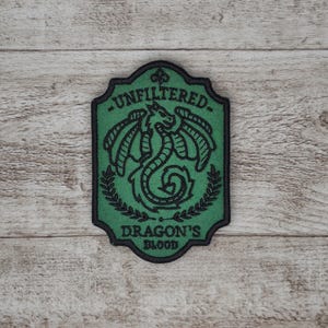May include: An embroidered patch in green and black, featuring the words "UNFILTERED" and "DRAGON'S BLOOD". The design includes a dragon illustration and a laurel wreath. The patch has a black border and a rectangular shape with a curved top.