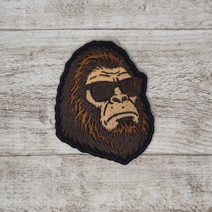 Peut inclure: Écusson brodé représentant un Bigfoot cool portant des lunettes de soleil. L'écusson a une couleur marron et beige avec une bordure noire. Le design représente un Sasquatch au visage sévère.