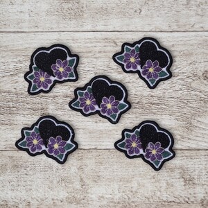 May include: Five embroidered patches. Each patch features a black heart shape with purple flowers and green leaves, set against a light wood grain background. Each patch has a white border.