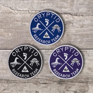 May include: Three circular embroidered patches featuring the text "CRYPTID RESEARCH TEAM" around the border. Each patch displays white icons of a footprint, a UFO, an eye in a triangle, and a sea creature arranged in a cross pattern on blue, black, and purple backgrounds.