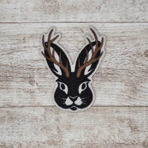 May include: Embroidered patch featuring a black rabbit head with brown antlers. The rabbit has white accents around the eyes, nose, and mouth. The patch is outlined in white and is likely designed to be ironed or sewn onto fabric.