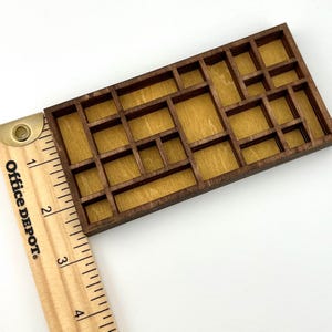 May include: A rectangular wooden grid display with multiple compartments of varying sizes, featuring a dark brown frame and a light tan backing. A wooden ruler is positioned along the left side, showing the object is approximately 4 inches long.