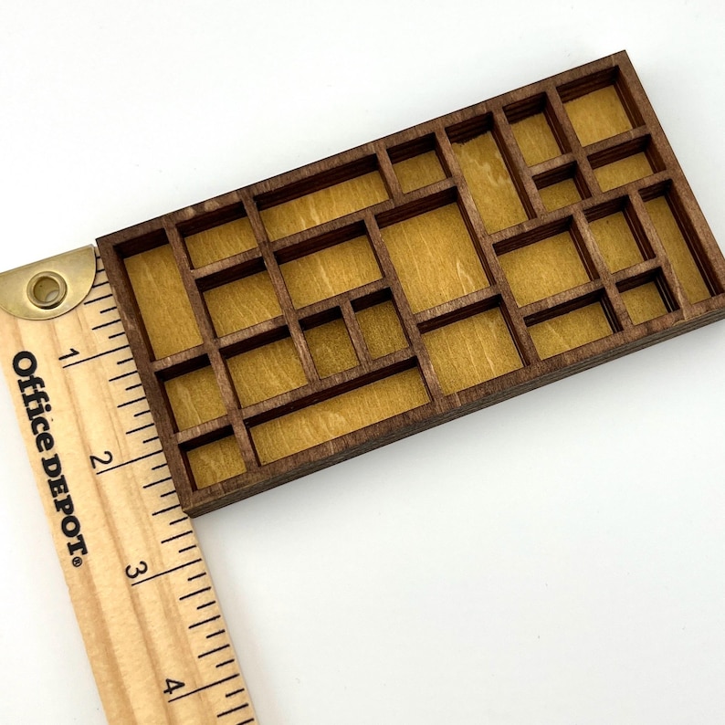 May include: A rectangular wooden grid display with multiple compartments of varying sizes, featuring a dark brown frame and a light tan backing. A wooden ruler is positioned along the left side, showing the object is approximately 4 inches long.