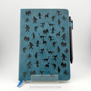 May include: Teal-colored notebook with a black pen. The notebook cover is decorated with black silhouettes of various mythical creatures. A blue and white ribbon bookmark is visible. The text "EXTRAKITSCH.COM" is printed at the bottom.