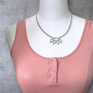 May include: A silver chain necklace with a silver, rhinestone-encrusted, knuckle duster pendant. The necklace is on a mannequin wearing a pink ribbed tank top.