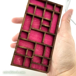 May include: A rectangular wooden display tray featuring a grid of various sized compartments with a vibrant hot pink felt lining. The dark brown wooden frame creates a structured geometric pattern of open cubbies for organizing small items.
