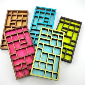 May include: Five rectangular wooden organizers with grid-like compartments. Each frame features a distinct color backing: natural wood, bright pink, turquoise, light blue, and neon yellow. The dividers are made of dark wood, creating a geometric pattern.