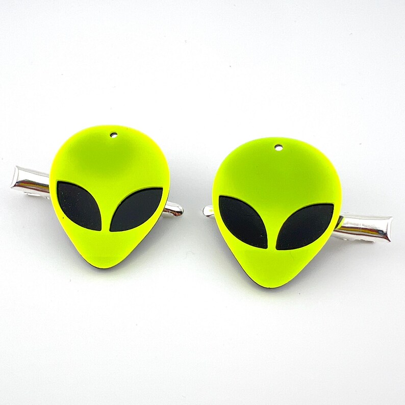 Op de afbeelding: Twee groene alien-hoofd haarclips met zwarte ogen. De clips zijn gemaakt van plastic en hebben een zilveren metalen clip aan de achterkant.