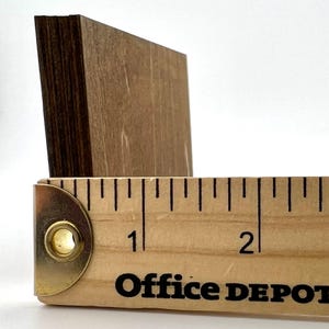 May include: A rectangular piece of light brown wood with a dark brown layered edge, positioned behind a wooden ruler that shows the object is approximately 1.5 inches wide. The ruler features black markings and the text Office DEPOT.