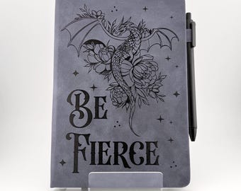 May include: Gray journal with a dragon and floral design, the words "BE FIERCE" are printed on the cover. A black pen is attached to the side. The journal is displayed on a clear stand.